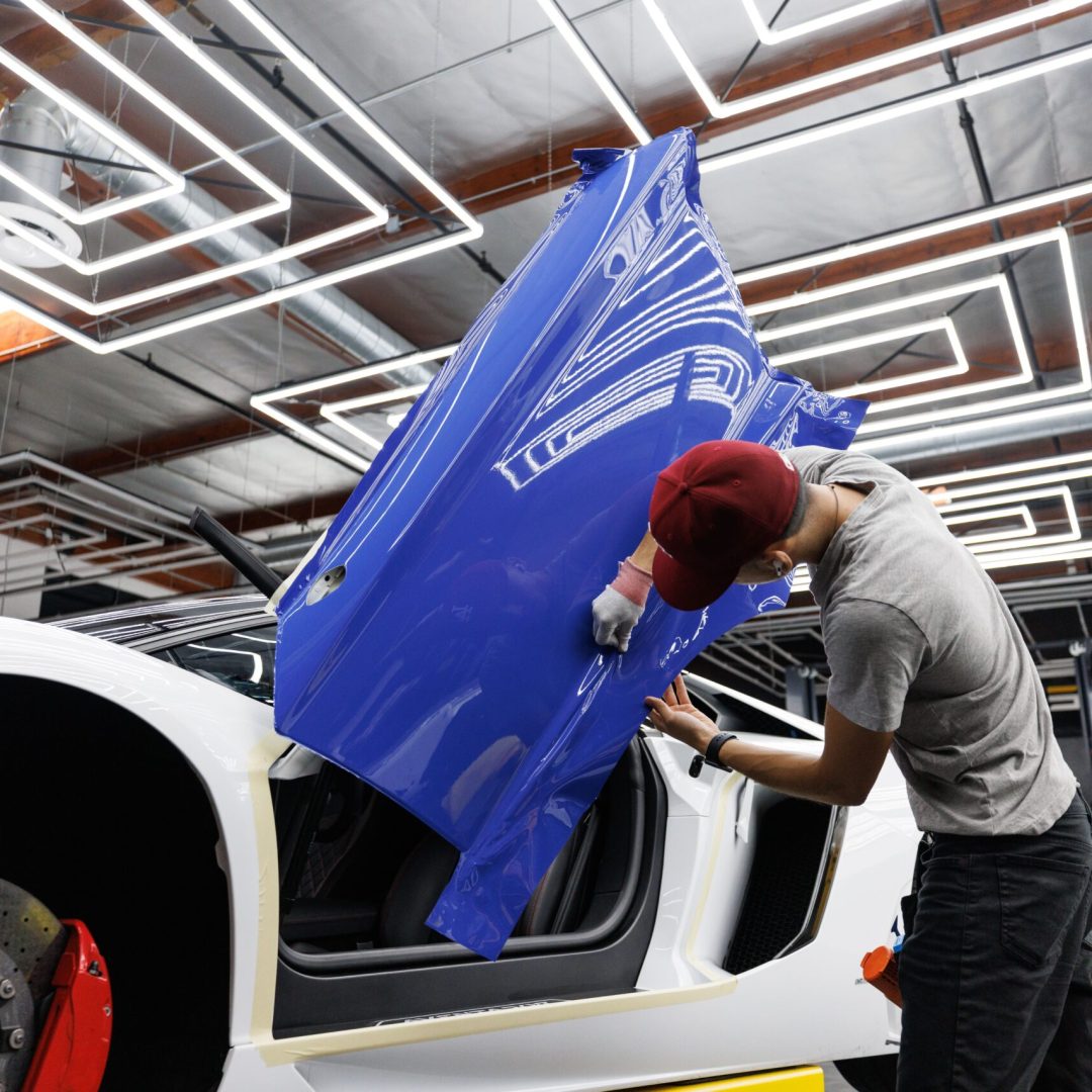 Auto mechanic applying blue vinyl wrap to a car door in a modern garage.