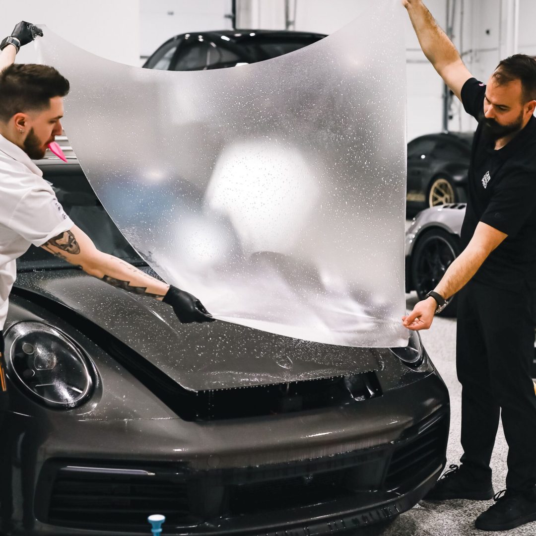 Two technicians applying a protective film on a sleek sports car in a garage setting.