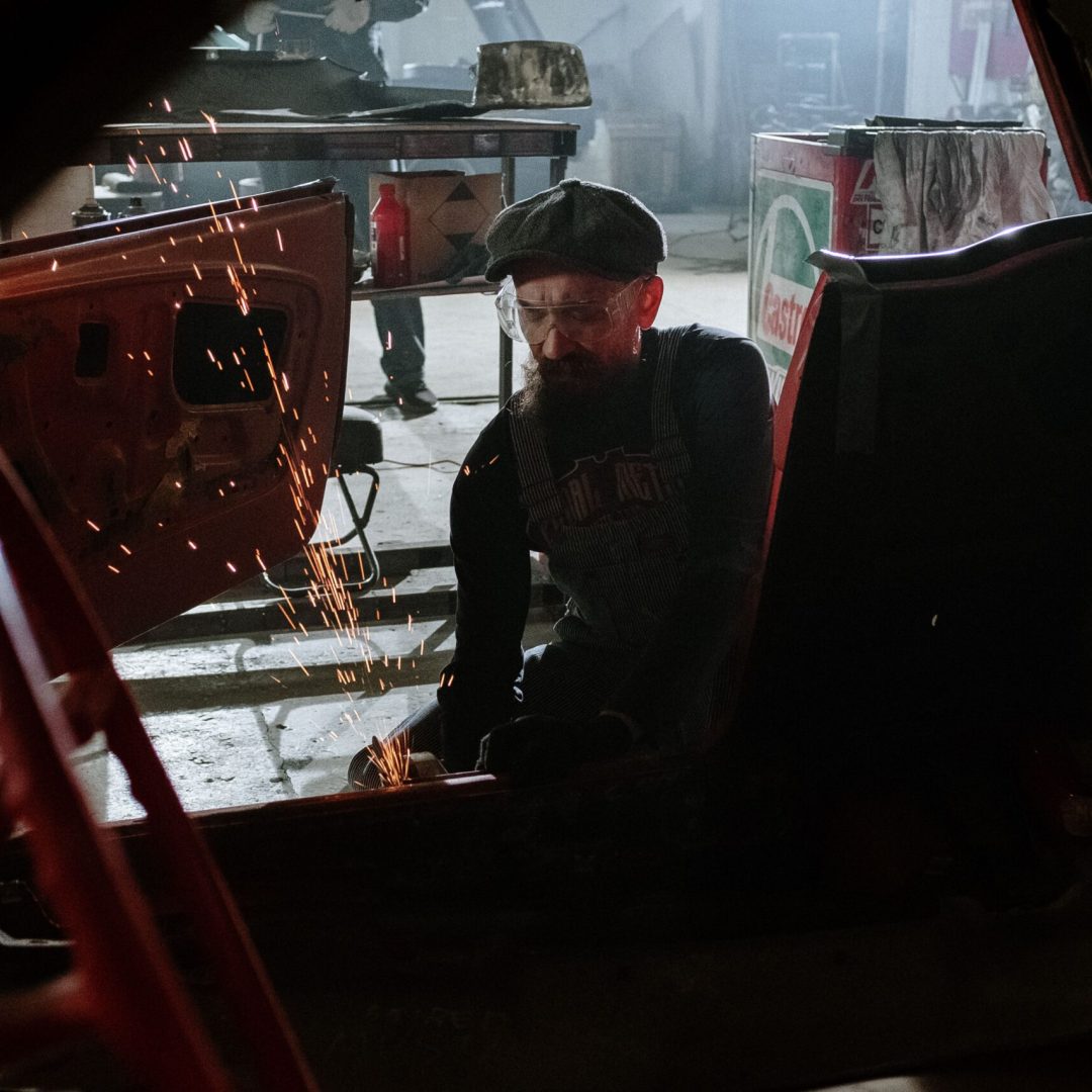 A mechanic welding inside an auto repair shop with flying sparks, focused on car restoration work.